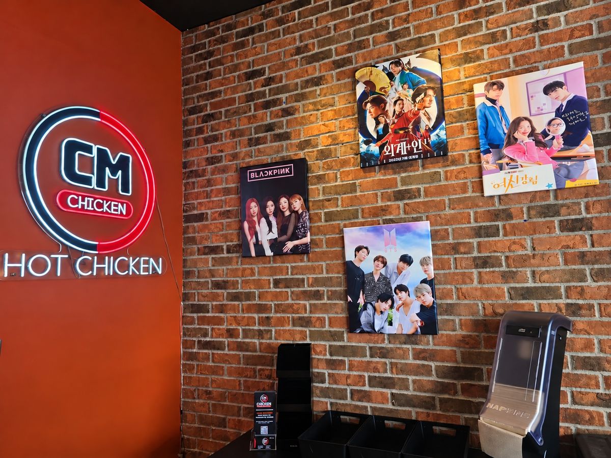 Neon CM logo and Hot Chicken sign on an orange wall beside K-pop, Korean film, and TV drama posters